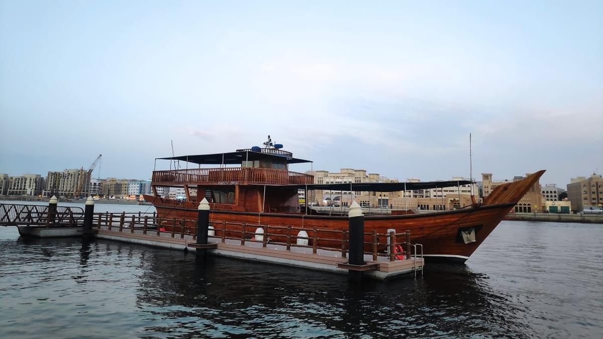 Traditional wooden dhow boat docked at Dubai Creek during daylight, offering visitor information and boarding details.