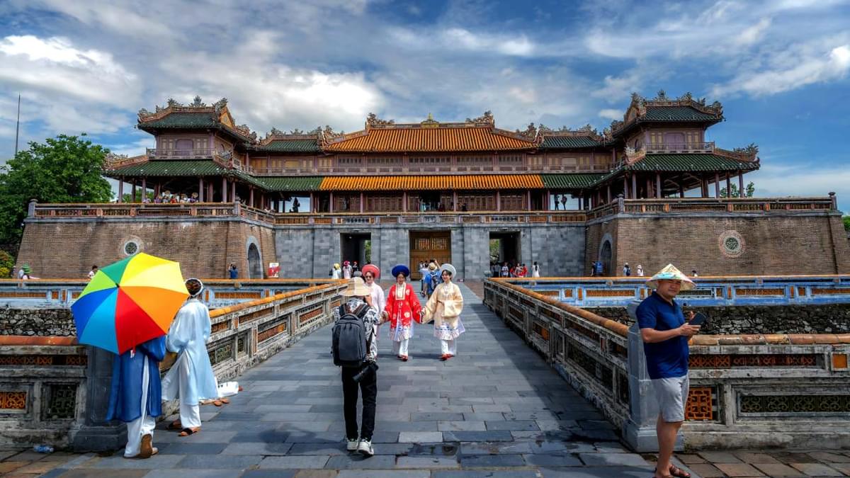 Tourists visiting the Imperial City of Hue in Vietnam, with traditional costumes, colorful umbrellas, and historic architecture under a partly cloudy sky.