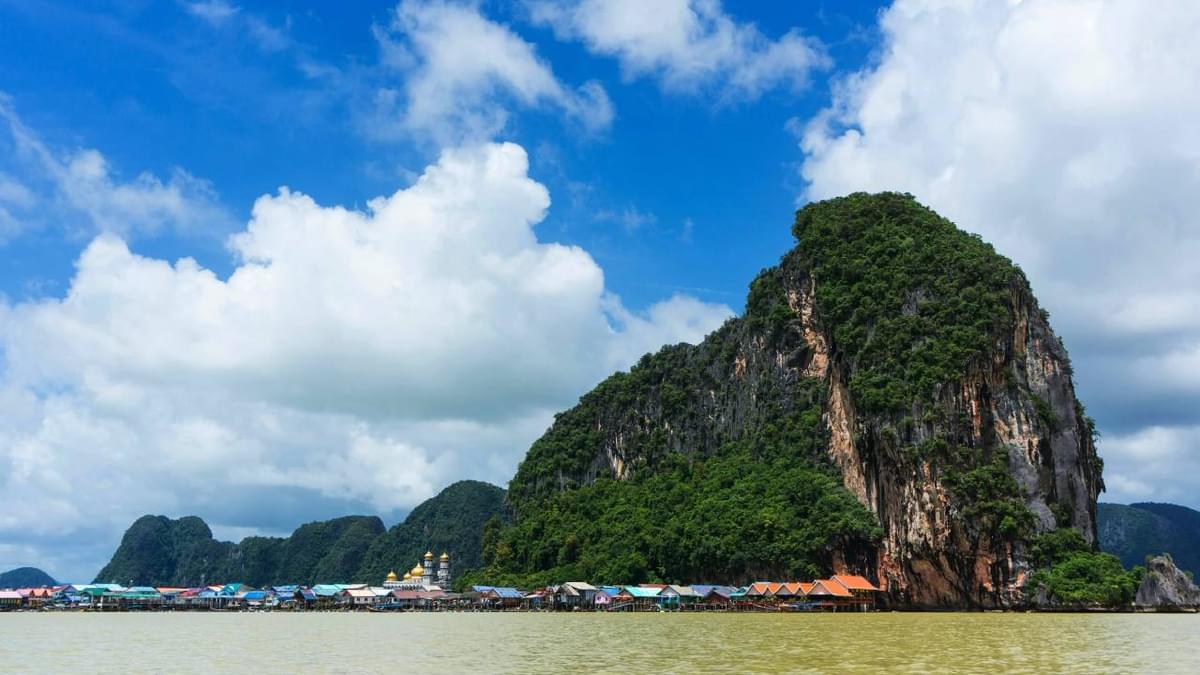 Scenic view of a floating village with colorful rooftops and a mosque, set against a towering limestone cliff and blue sky in Phang Nga Bay, Thailand.