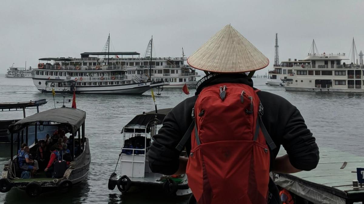 Traveler with a red backpack and traditional conical hat looking at cruise boats in Halong Bay, Vietnam, on a cloudy day.
